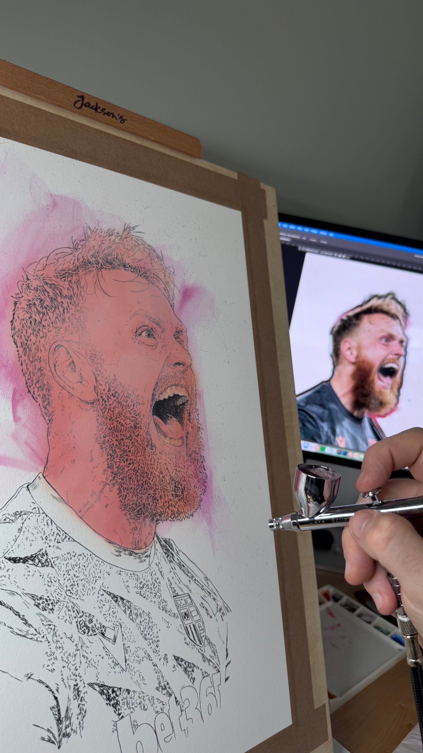 Close-up of a work-in-progress portrait of Viktor Johansson beside its reference image, with an airbrush held in the foreground.